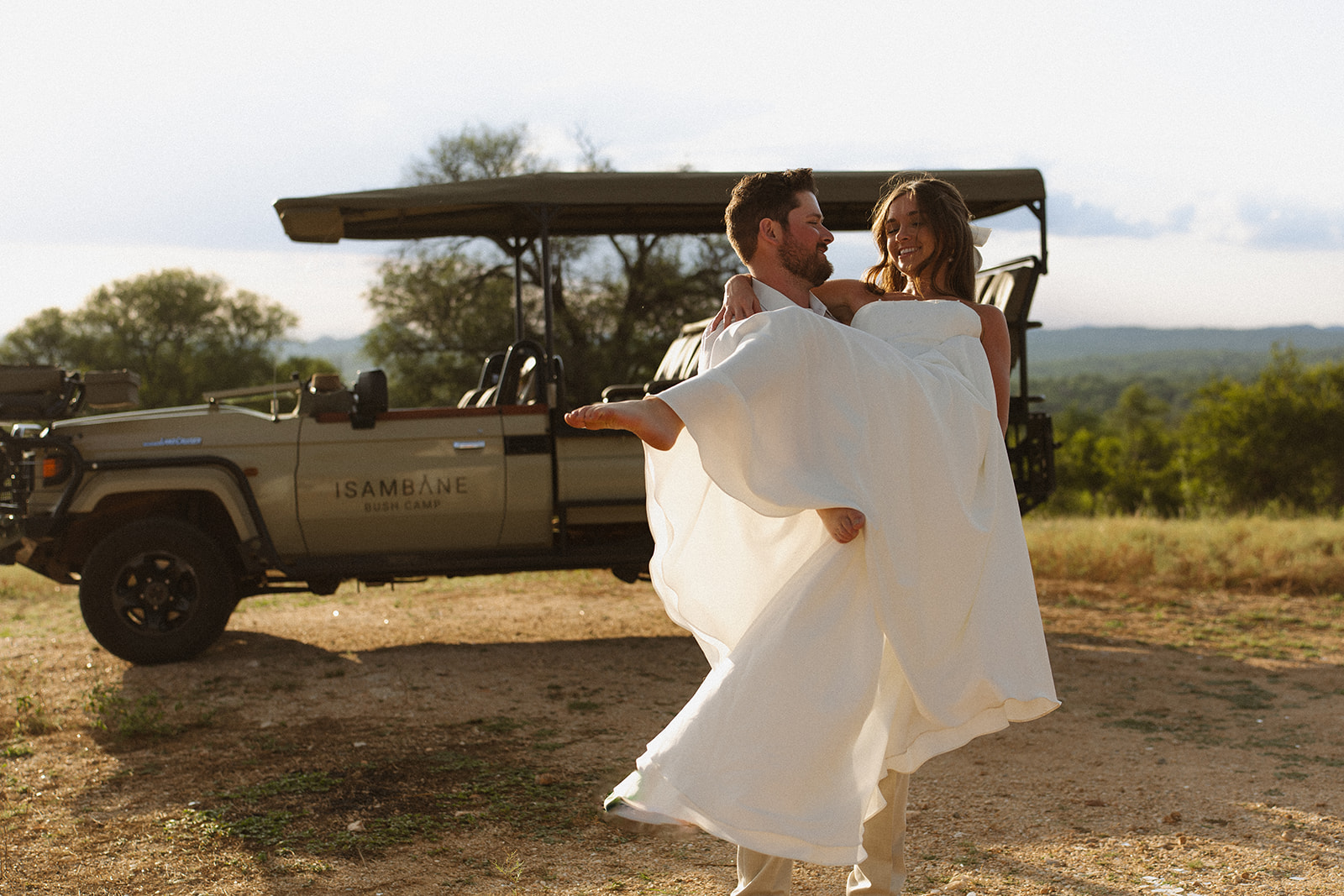 Groom lifting bride in front of safari vehicle with flowing dress and golden light during their wedding day
