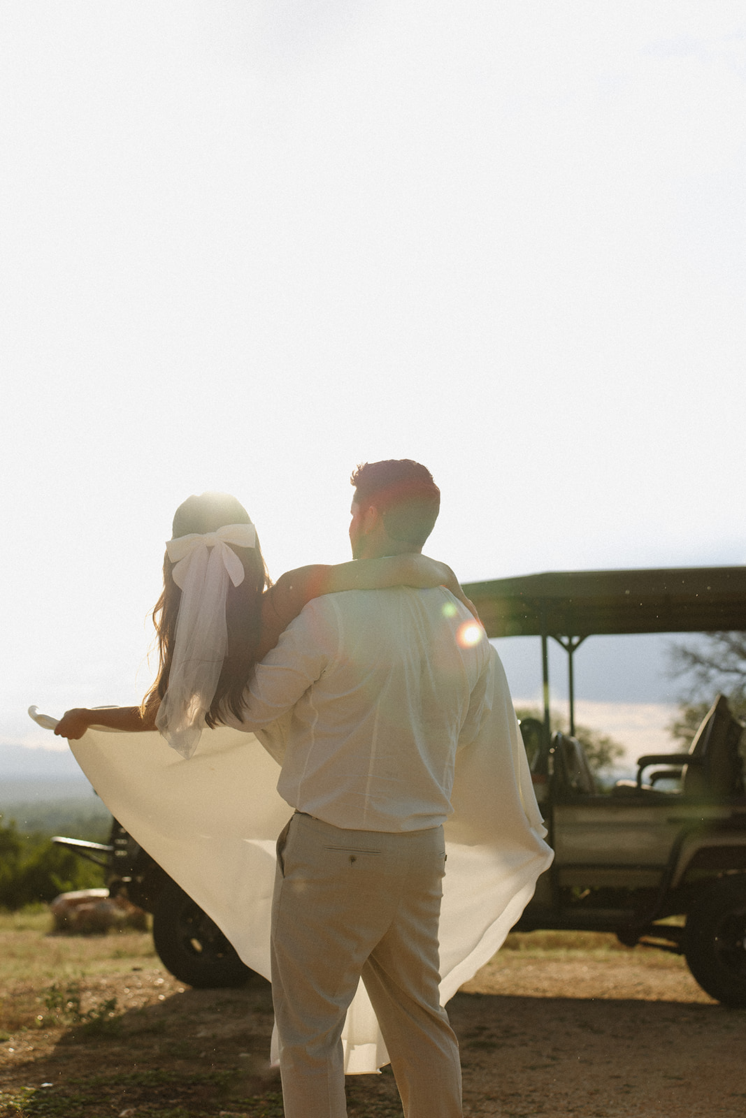 Groom lifting bride with flowing dress in golden backlight beside safari vehicle during a safari wedding packages experience
