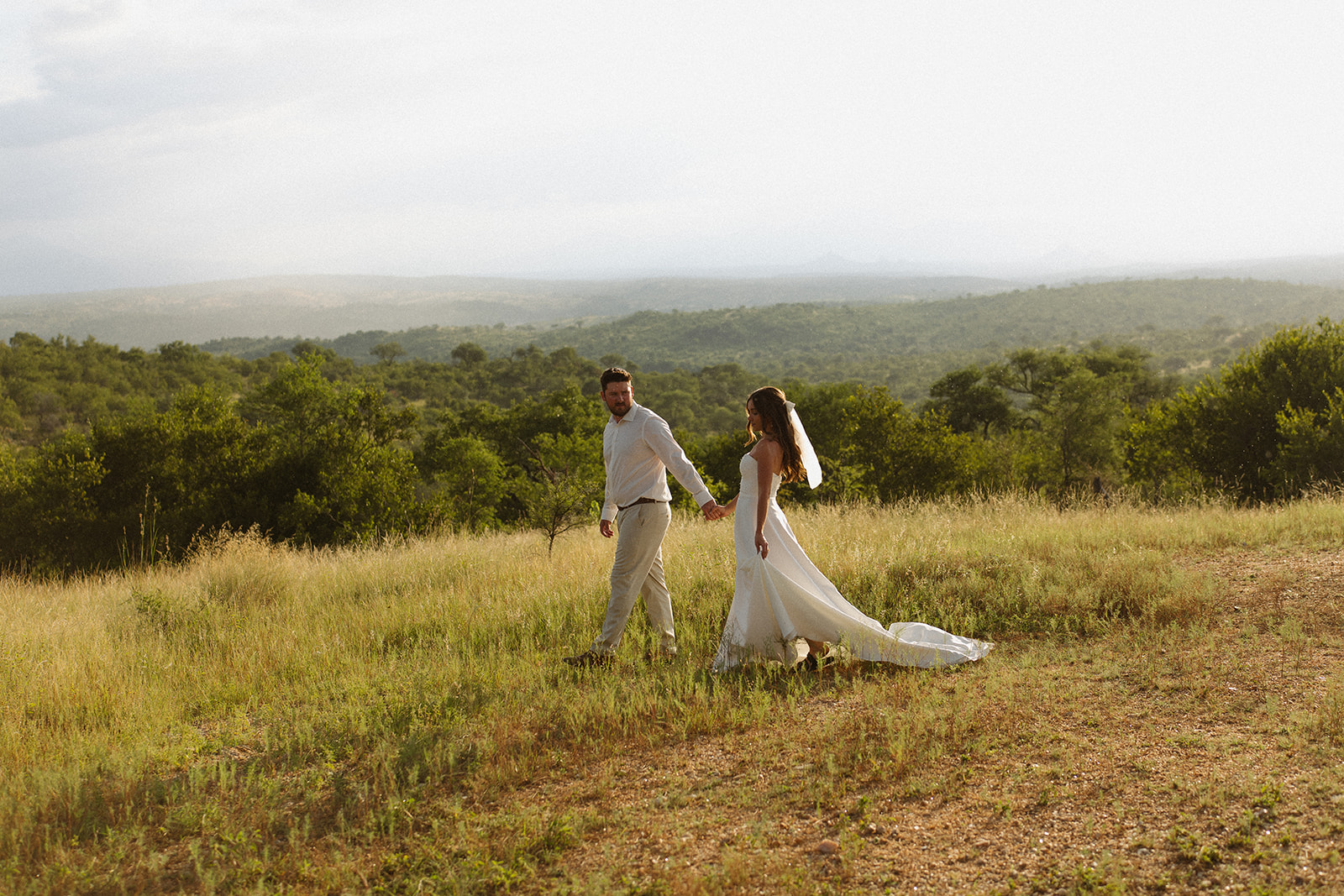 Bride and groom walking hand in hand through grassy African landscape with scenic mountain views on their wedding day
