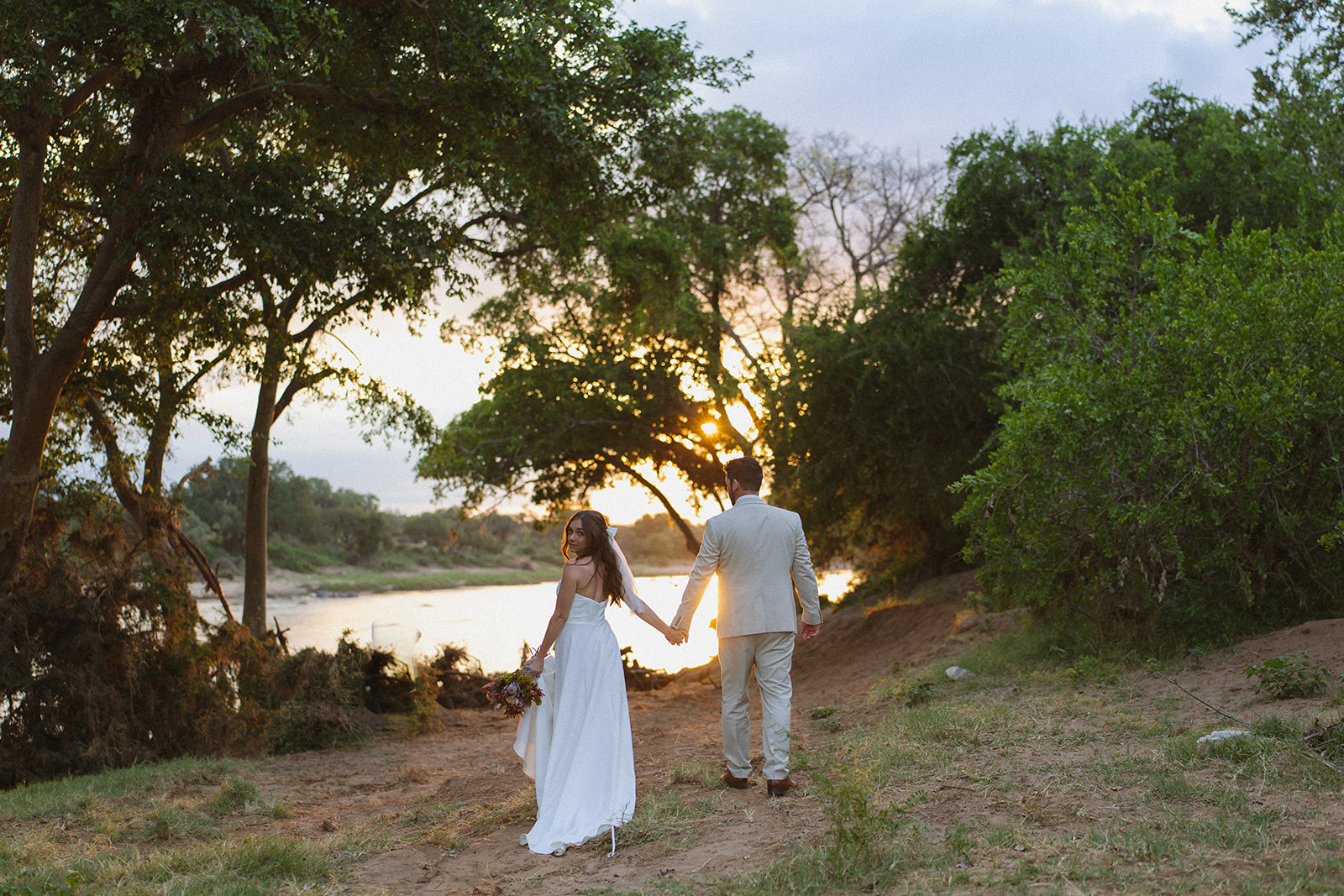 Bride and groom walking hand in hand along a river at sunset surrounded by lush trees during a safari wedding packages experience
