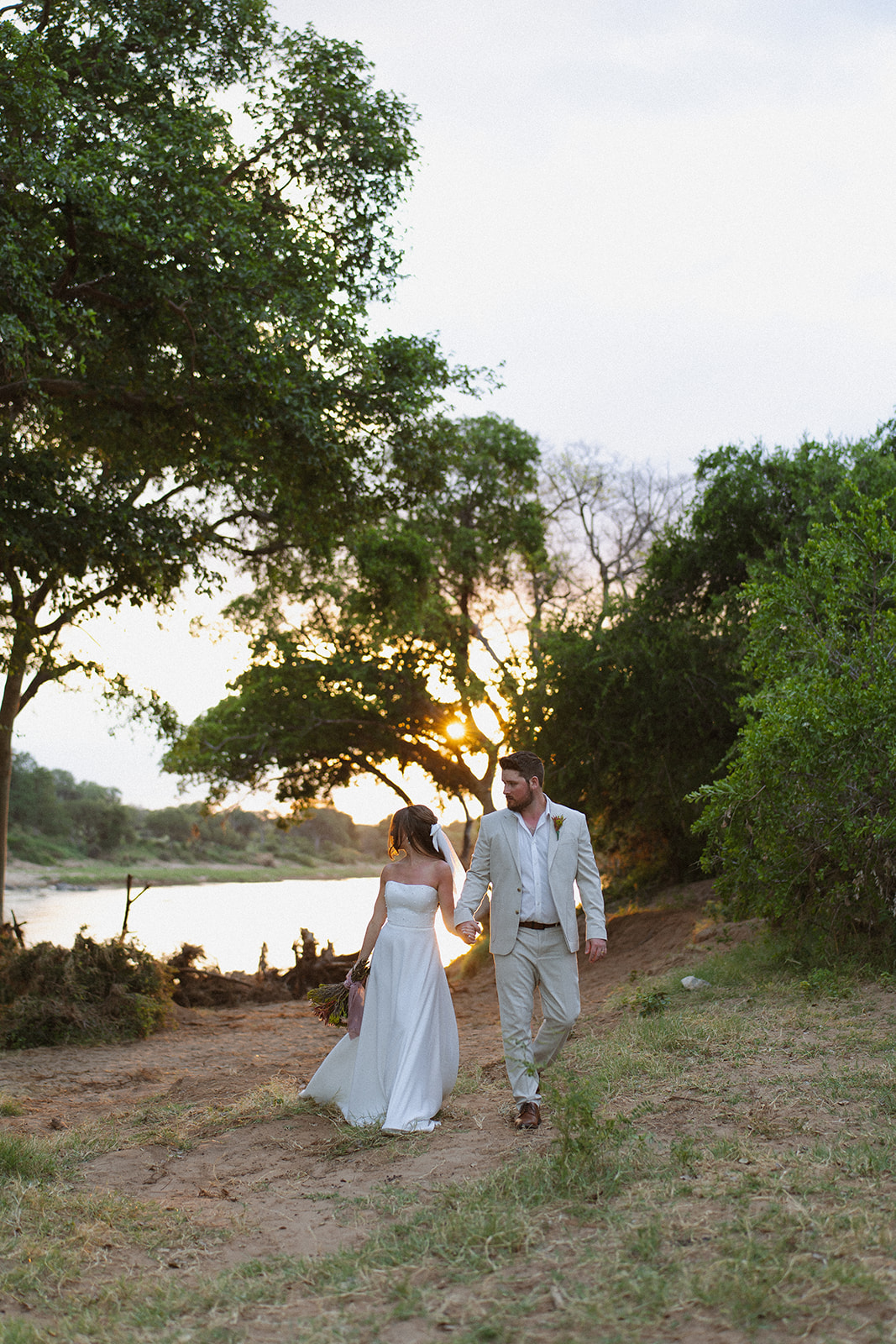 Bride and groom strolling by the water at golden hour with soft light filtering through trees on their wedding day
