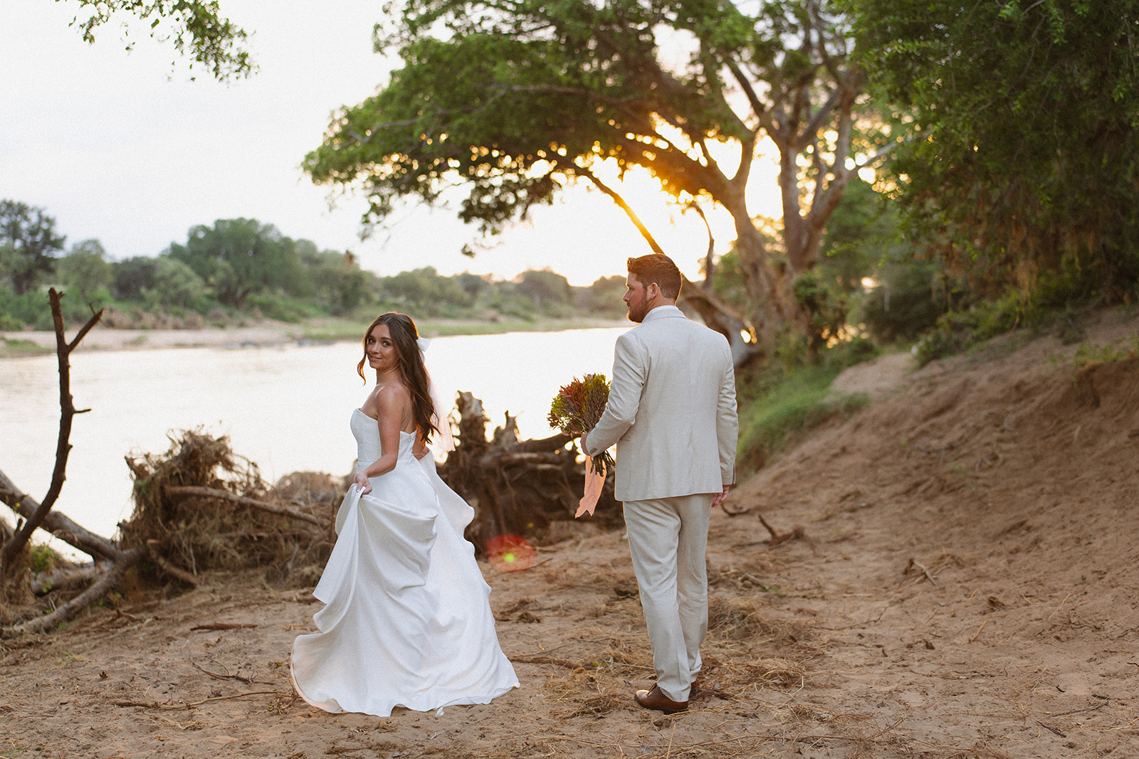 Bride and groom walking along the riverbank at sunset with bouquet and flowing dress on their wedding day
