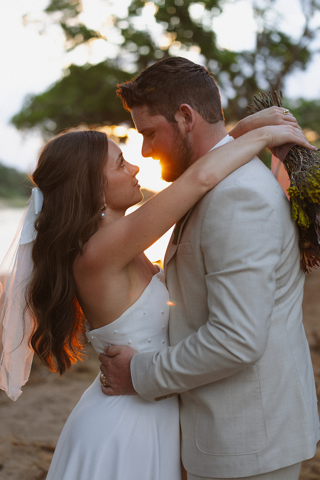 Bride and groom embracing in golden sunset light by the river with bouquet during their wedding day
