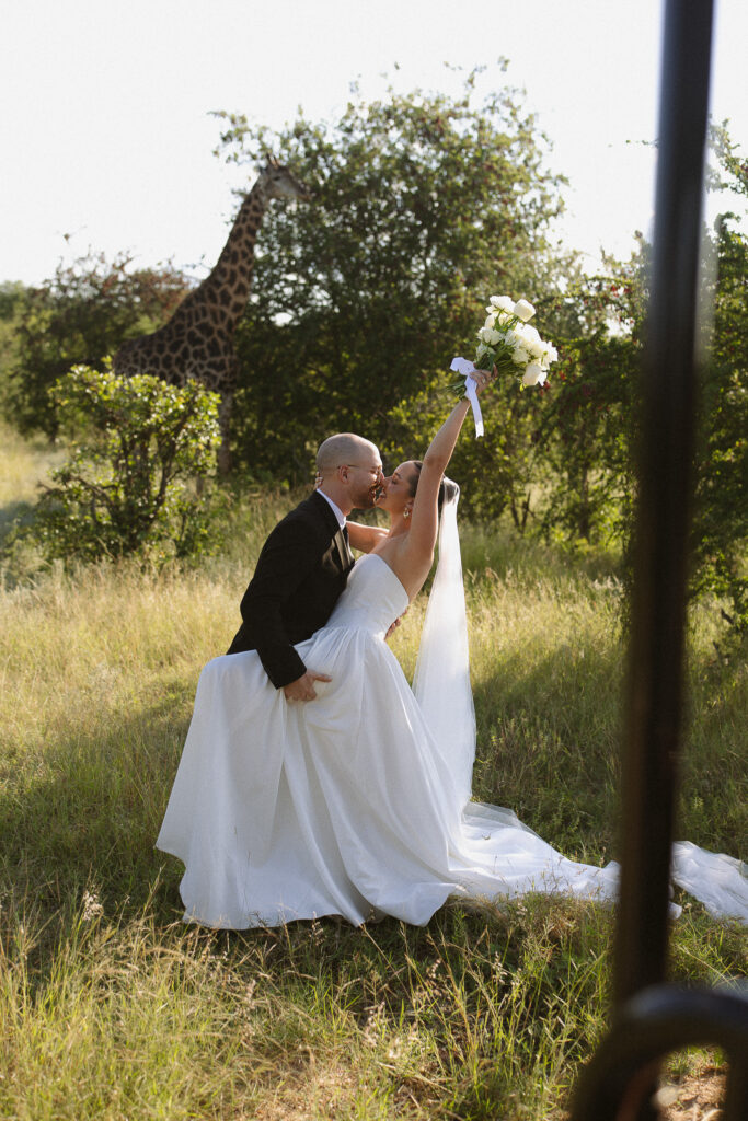 Bride and groom celebrating in the bush with a giraffe behind them during their South Africa safari wedding