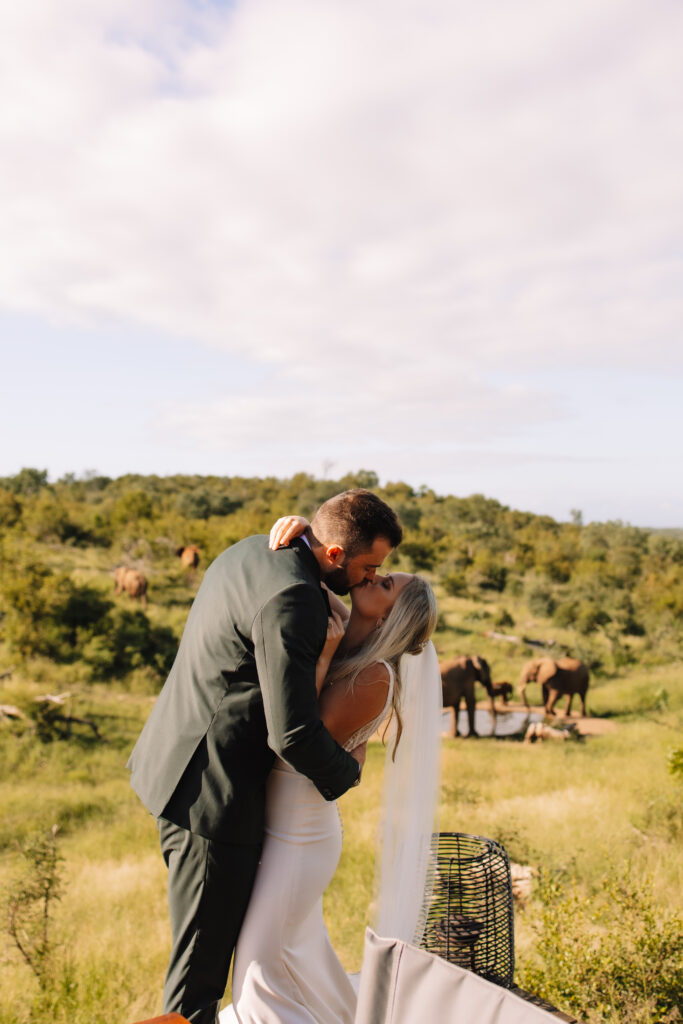 Bride and groom kissing during their safari wedding with elephants gathered at a waterhole behind them