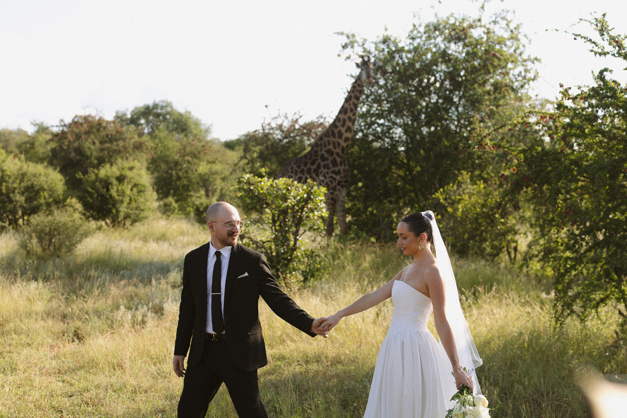 Bride and groom holding hands with a giraffe behind them during a safari wedding in South Africa