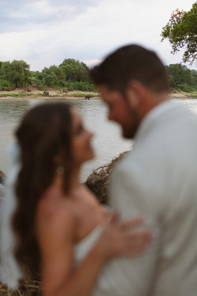 Couple embracing beside the river with elephants in the distance during their South Africa safari vow renewal