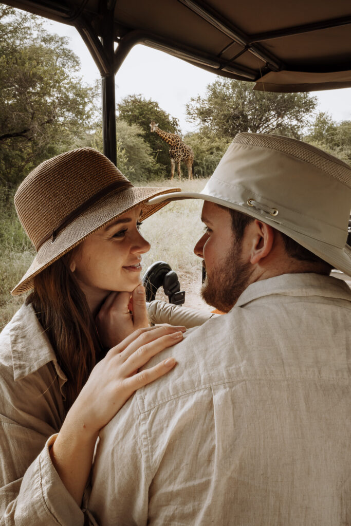 Couple sitting together in a safari vehicle with a giraffe behind them in Sabi Sands